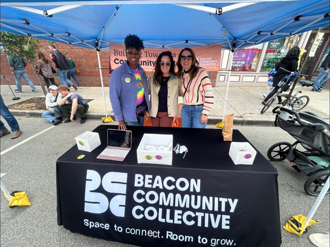 Three people standing behind the Beacon Community Collective table under a blue tent, the tablecloth reading "Space to connect. Room to grow."