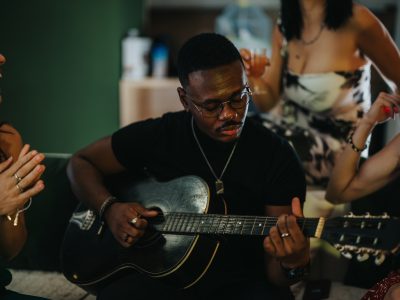 A man playing guitar while friends clap and enjoy a lively atmosphere at a cheerful gathering, suggesting fun and community around music and happiness.